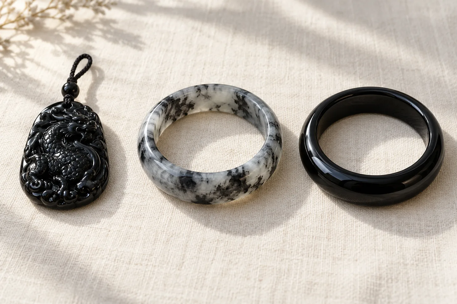 Three dark pieces in a row on ivory linen: a carved black jadeite pendant, a grey-white bangle with black patches, and a glossy, evenly black bangle