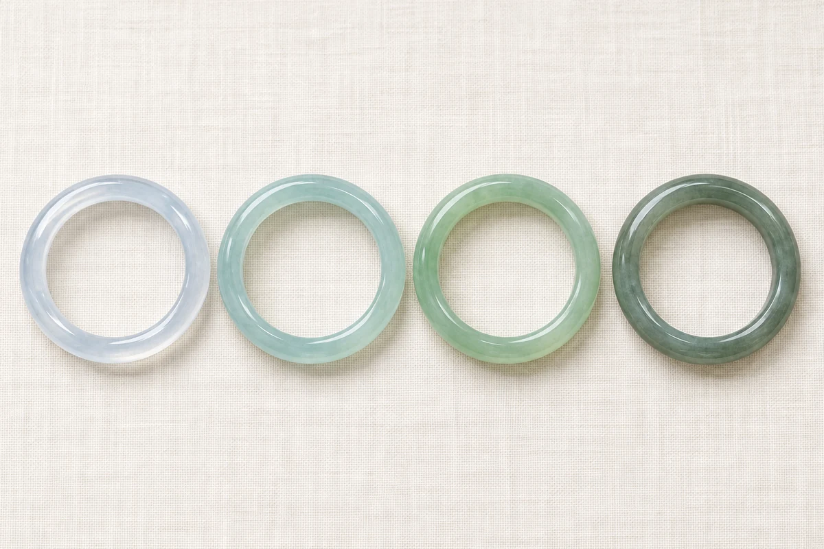 Four jadeite bangles in a row on ivory linen in soft daylight, left to right: a very transparent bangle with a pale cool blue tone, a transparent bangle with an even pale blue-green, a translucent bangle with an even pale green, and a darker, greyish green bangle with an oily sheen