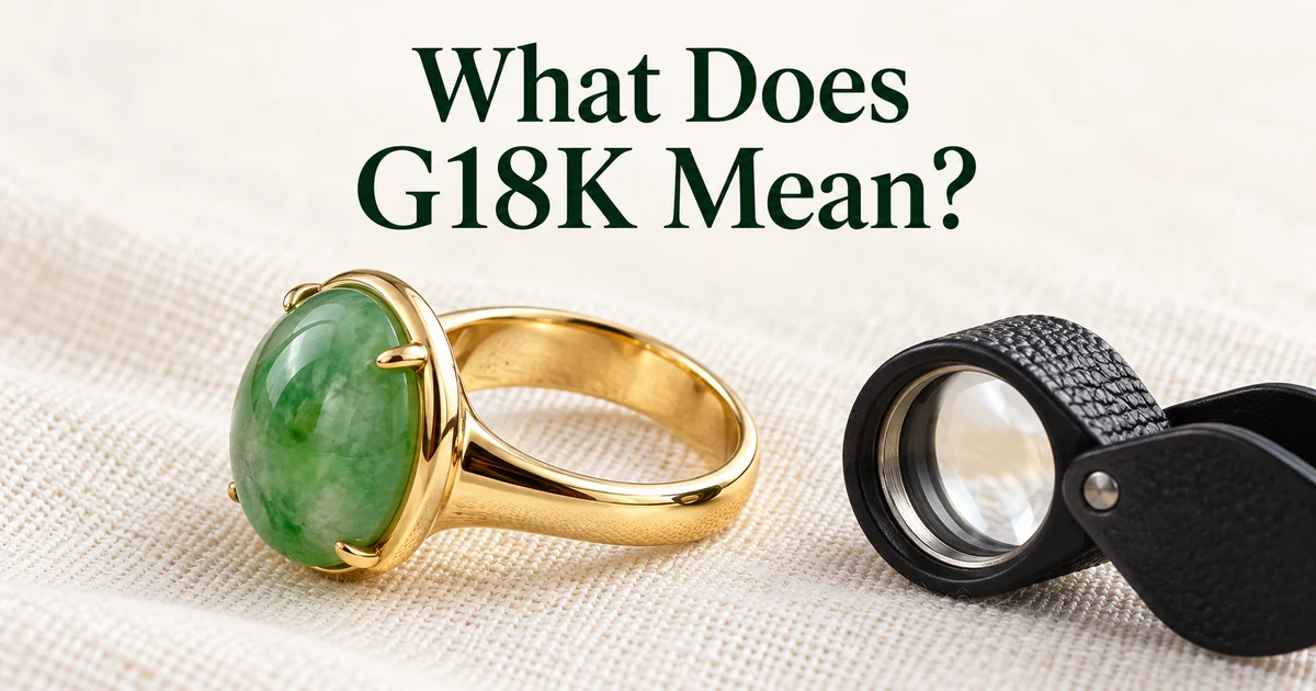 A green jadeite cabochon ring in yellow gold lying on its side on ivory linen so the inside of the band shows, next to a jeweler's loupe, in soft daylight