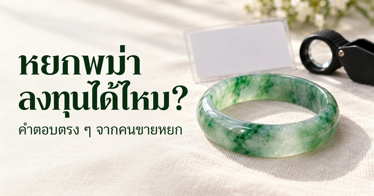 กำไลหยกพม่าสีเขียวใสวางชัดอยู่ด้านหน้าบนผ้าลินินสีงาช้าง ด้านหลังเป็นการ์ดใบรับรองว่างเปล่าและแว่นขยายส่องพลอยที่เบลออยู่ ในแสงธรรมชาติอ่อน ๆ