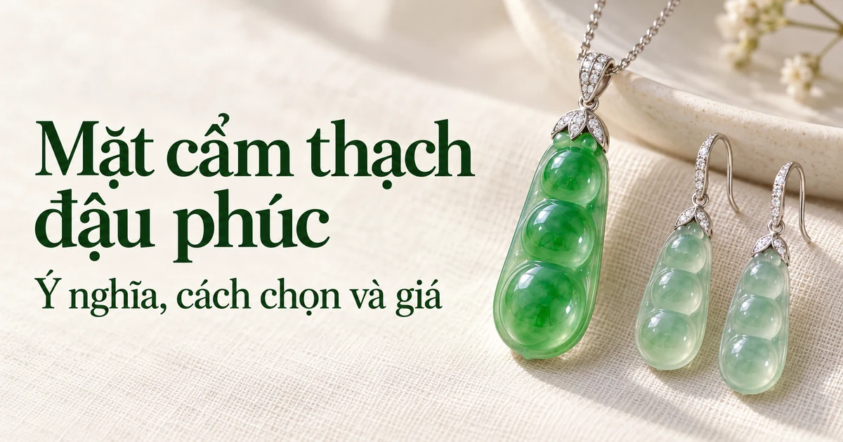 Một mặt đậu phúc phỉ thúy cao băng xanh lục tươi có chụp vàng trắng trên dây chuyền vàng trắng mảnh, nằm trên nền vải lanh màu ngà cạnh một đôi bông tai đậu phúc băng chủng màu nhạt, dưới ánh sáng ban ngày dịu