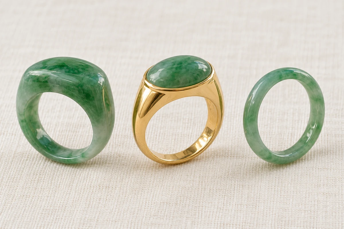 Three rings in a row on ivory linen, left to right: an all-jadeite green saddle ring with a raised saddle-shaped top, a yellow-gold saddle ring set with a green jadeite cabochon, and a plain green jadeite band for comparison