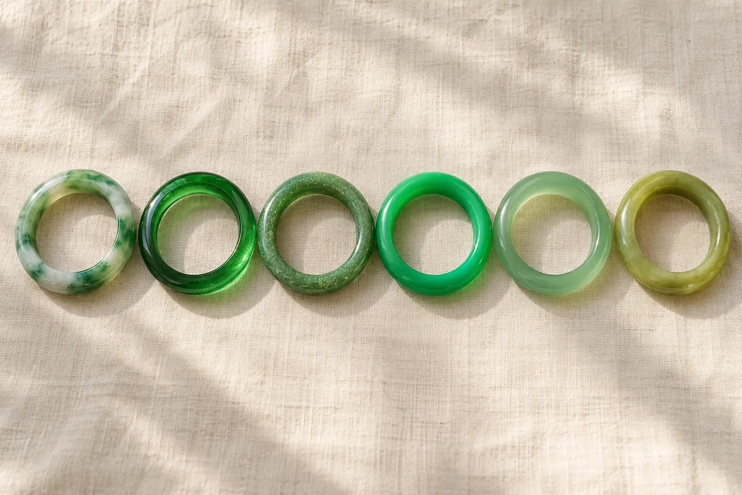 Six green bangles in a row on ivory linen: natural jadeite, glass, aventurine quartz, dyed quartzite, green chalcedony and serpentine
