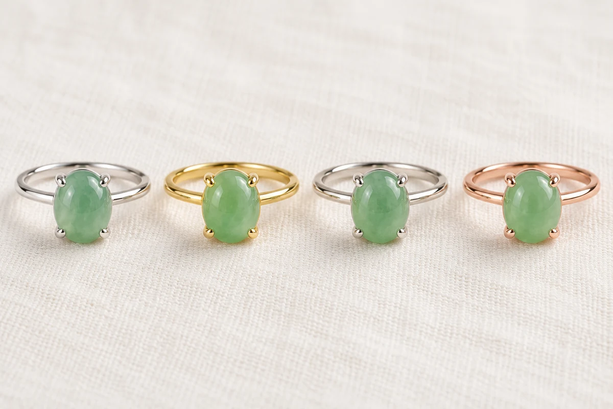 Four rings in a row on ivory linen, each set with a similar light green jadeite cabochon: a sterling silver ring, an 18K yellow gold ring, an 18K white gold ring and an 18K rose gold ring