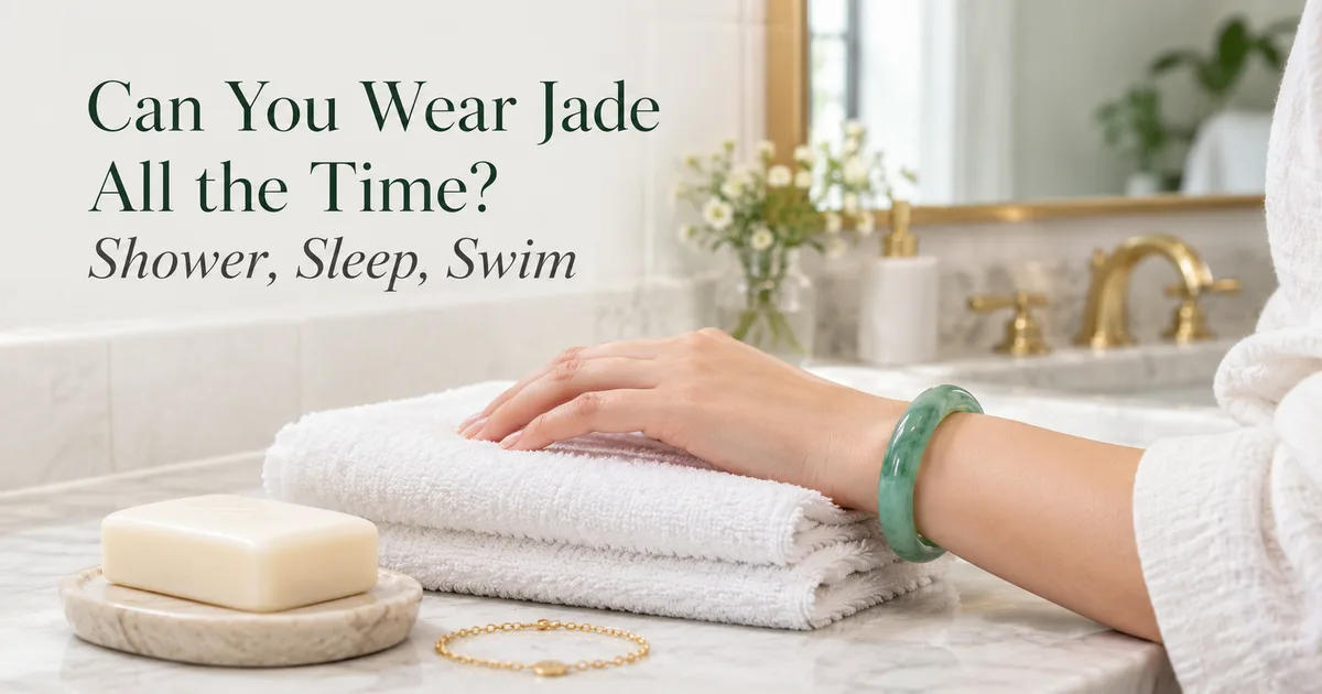 A green jadeite bangle on a wrist resting on a folded white towel beside a bathroom sink, a bar of soap and a gold bracelet nearby, with the title Can You Wear Jade All the Time? Shower, Sleep, Swim
